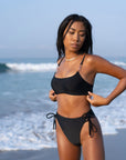 Woman on a beach wearing the Azalea Bottom / Black, a high-waisted, cheeky swimsuit with side lacing and rose gold hardware.