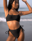 Woman wearing Azalea Bottom in black, high-waisted with lace-up sides, showcasing cheeky bottom coverage on a beach.