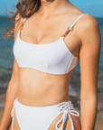 A woman wearing the Azalea Bottom in white, featuring high waisted, high leg cut, cheeky coverage, and adjustable lace-up sides with rose gold hardware.