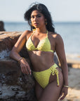 Woman in yellow Azalea Bottom; high-waisted, lace-up sides, cheeky coverage, rose gold hardware, seamless fit.
