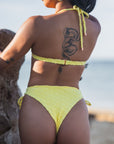 Woman wearing Azalea Bottom in yellow, a high-waisted, cheeky lace-up bikini bottom with rose gold hardware and seamless design, showcasing a dragon tattoo on her back.