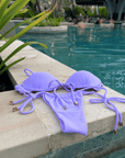 Jasmine Bottom / Lilac swimsuit with adjustable drawstring and cheeky cut, displayed on a ledge beside a pool.