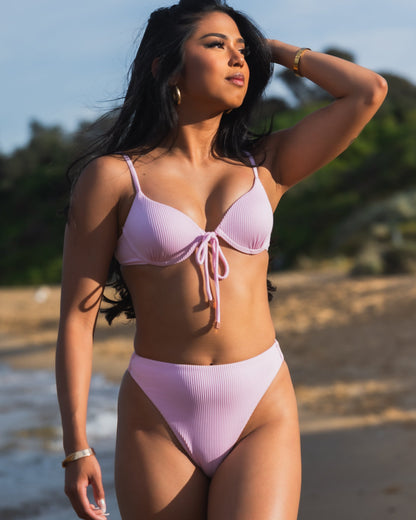 Woman wearing Blossom Bottom / Pink high-waisted bikini on a beach, showcasing cheeky bottom coverage and rose gold hardware.